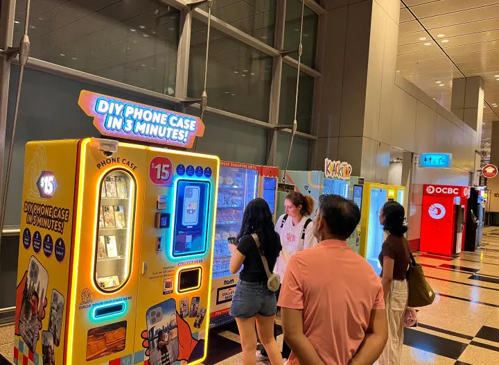 A modern DIY phone case vending machine in a busy shopping mall atrium, showing customers interacting with the touchscreen
